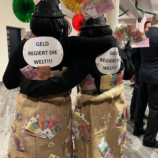 Zwei Personen in Kostüm, jeweils mit einem mit Geld gefüllten Sack aus Jute, stehen Rücken an Rücken. Sie tragen Schilder mit der Aufschrift 'GELD REGIERT DIE WELT!!'. Ein Mann in Anzug steht hinter ihnen.