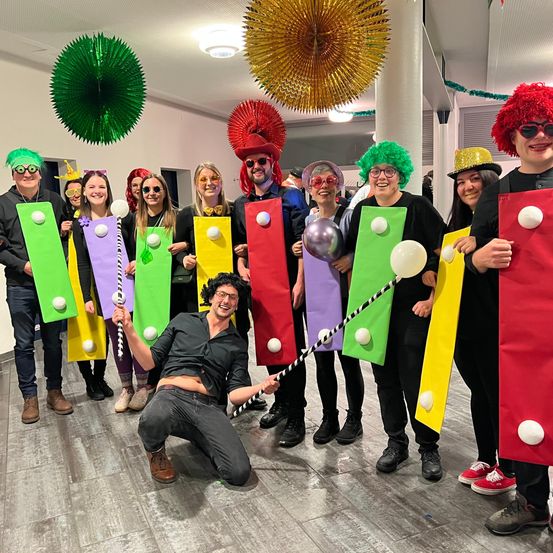 Eine Gruppe von Menschen in bunten Kostümen und mit Requisiten versammelt sich in einem Raum, einige knien, andere stehen, mit einer festlichen Atmosphäre und Dekorationen.