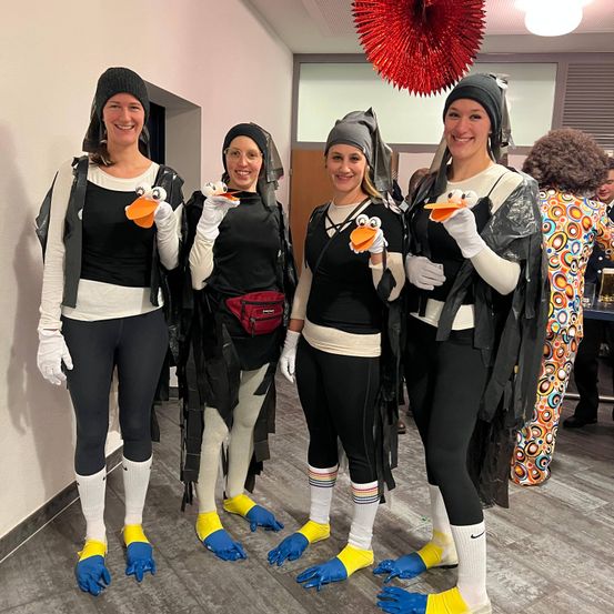 Vier Frauen in Pinguinkostümen, die Schnabel-Requisiten halten, stehen in einer Reihe. Sie tragen Handschuhe, Socken und gelbe Füße, eine trägt eine rote Tasche. Dahinter hängt eine rote Dekoration von der Decke.