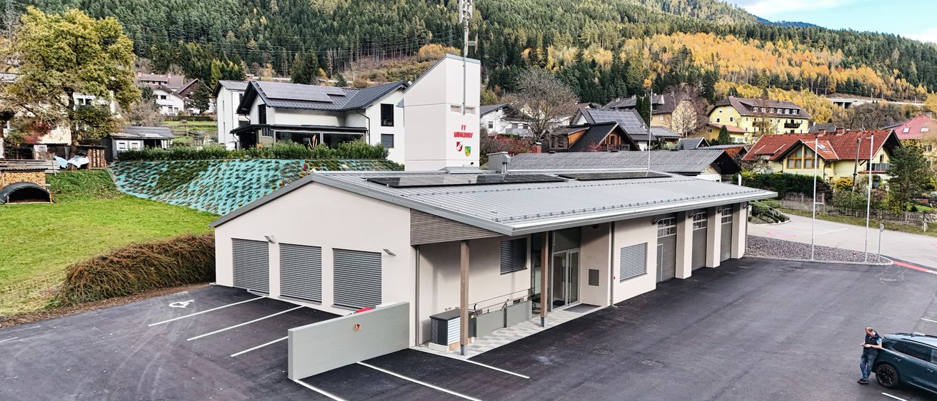 Ein Luftbild einer modernen Feuerwache mit Solarmodulen auf dem Dach, umgeben von Häusern und Bergen im Hintergrund.