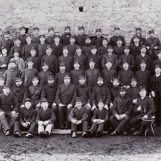 Eine Gruppe von Männern, wahrscheinlich eine militärische Einheit, in Uniform gekleidet, posiert für ein Schwarz-Weiß-Foto. Sie sitzen und stehen vor einer Steinmauer.