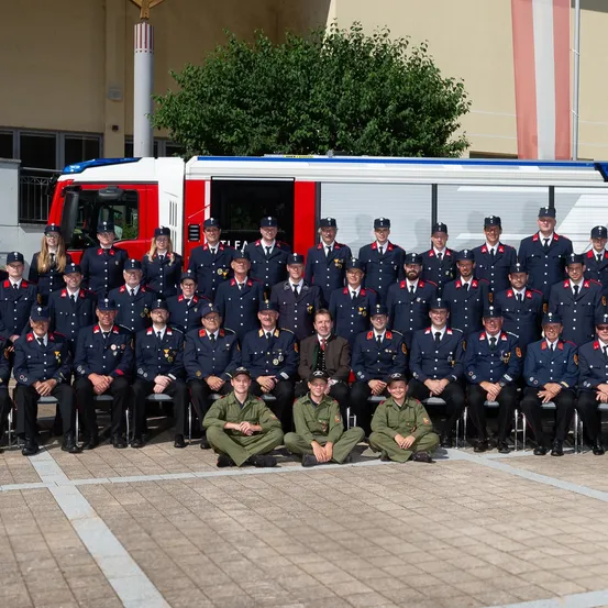 Eine Gruppe von Feuerwehrleuten, sowohl stehend als auch sitzend, posiert für ein Foto vor einem Feuerwehrwagen.