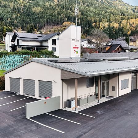 Eine Draufsicht auf ein modernes Gebäude mit Flachdach und Solarmodulen, umgeben von einem Parkplatz, Häusern und einem bewaldeten Berg im Hintergrund.