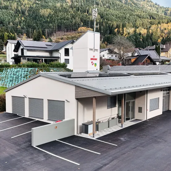 Eine Draufsicht auf ein modernes Gebäude mit Flachdach und Solarmodulen, umgeben von einem Parkplatz, Häusern und einem bewaldeten Berg im Hintergrund.
