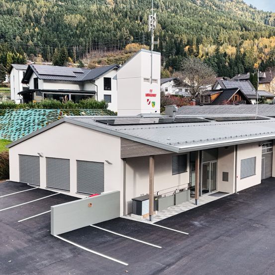 Eine Draufsicht auf ein modernes Gebäude mit Flachdach und Solarmodulen, umgeben von einem Parkplatz, Häusern und einem bewaldeten Berg im Hintergrund.