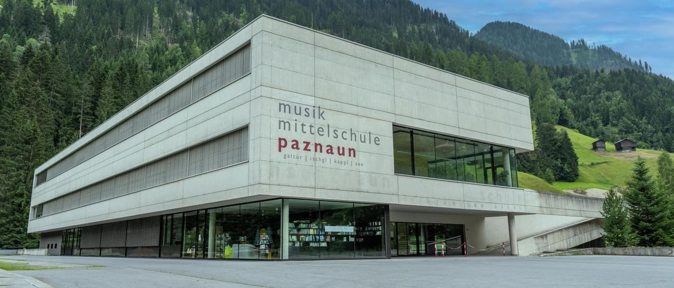 Ein modernes Gebäude mit der Aufschrift 'musik mittelschule paznaun' an der Wand, vor einer Bergkette mit üppigem Grün.