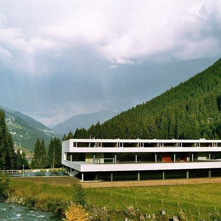 Ein modernes weißes Gebäude mit einem geschwungenen Dach steht in einem üppigen grünen Tal, umgeben von Bergen und einem Fluss.
