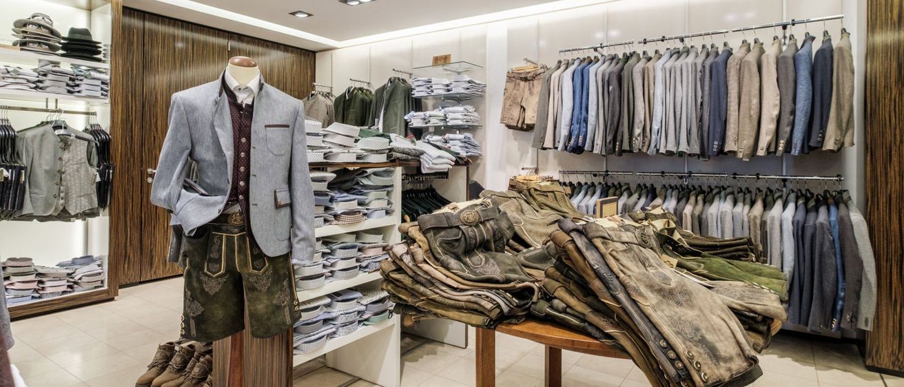 Ein Bekleidungsgeschäft zeigt eine Vielzahl von Artikeln. Ein Mannequin in einer Weste und Shorts steht in der Mitte. In der Nähe steht ein Tisch mit gefalteten Hosen. Die Wände sind mit hängenden Hemden gesäumt. Die Decke ist mit quadratischen Lichtern versehen.