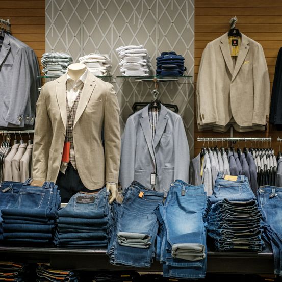 Ein Bekleidungsgeschäft zeigt eine Vielzahl von Herrenanzügen und Jeans. Auf einem schwarzen Displaytisch stehen in Anzügen gekleidete Mannequins, umgeben von ordentlich gefalteten Jeans. Der Laden verfügt über Holzwände mit Rautenmuster und Glasregalen mit gefalteten Hemden.