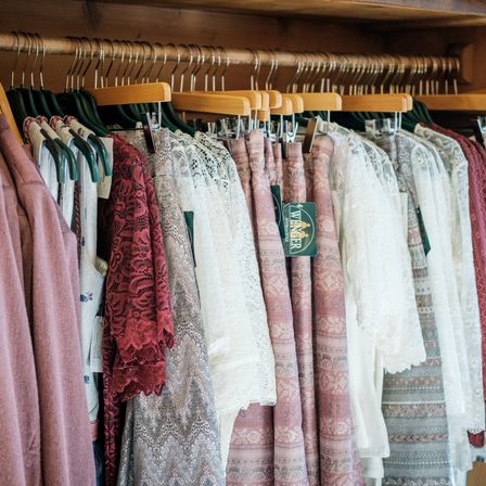 Ein Schrank voller verschiedener Damenbekleidung. Die Kleider hängen an Holzbügeln und umfassen Spitzen-, Blumen- und einfarbige Kleider.