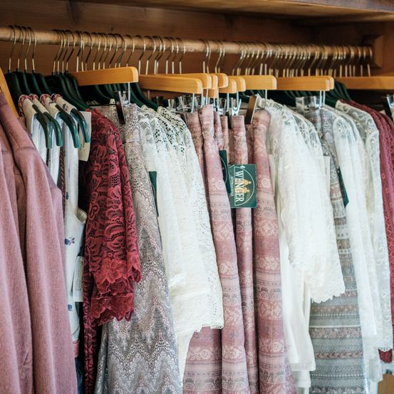 Ein Schrank voller verschiedener Damenbekleidung. Die Kleider hängen an Holzbügeln und umfassen Spitzen-, Blumen- und einfarbige Kleider.