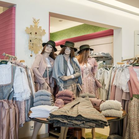 Drei in Pastellfarben gekleidete Mannequins befinden sich in einem Bekleidungsgeschäft, mit Garderobenständern für Pastellkleider und -blusen dahinter. Ein Tisch hat verschiedene gefaltete Kleidungsstücke darauf.