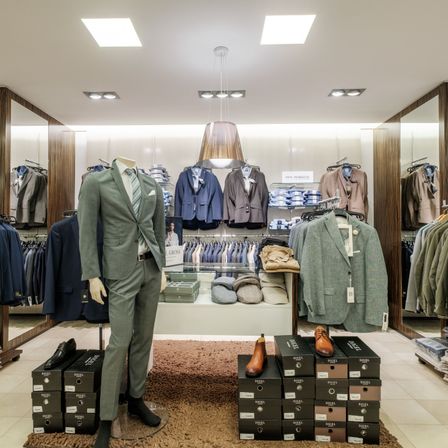 Ein Geschäft zeigt verschiedene Anzüge und Schuhe. Ein Dummy trägt einen grünen Anzug und schwarze Schuhe. Dahinter hängen Anzüge an Kleiderbügeln und Schuhe sind auf Kisten gestapelt. Ein brauner Teppich bedeckt den Boden.