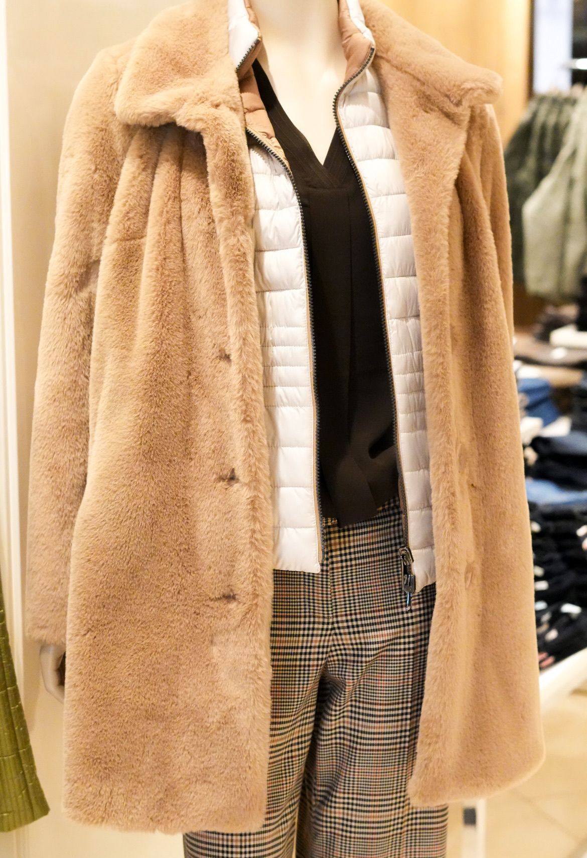 Ein Dummy zeigt einen hellbraunen Pelzmantel über einem schwarzen Oberteil und karierten Hosen, mit einer weißen, puffigen Jacke darunter.