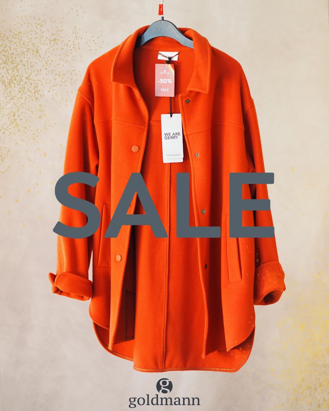 Ein orangefarbener Mantel mit einem Schild, das -50% SALE und WIR SIND GERRY steht. Der Mantel hängt an einem Kleiderbügel. Das Wort SALE ist in großen Buchstaben auf einem weißen Hintergrund geschrieben.