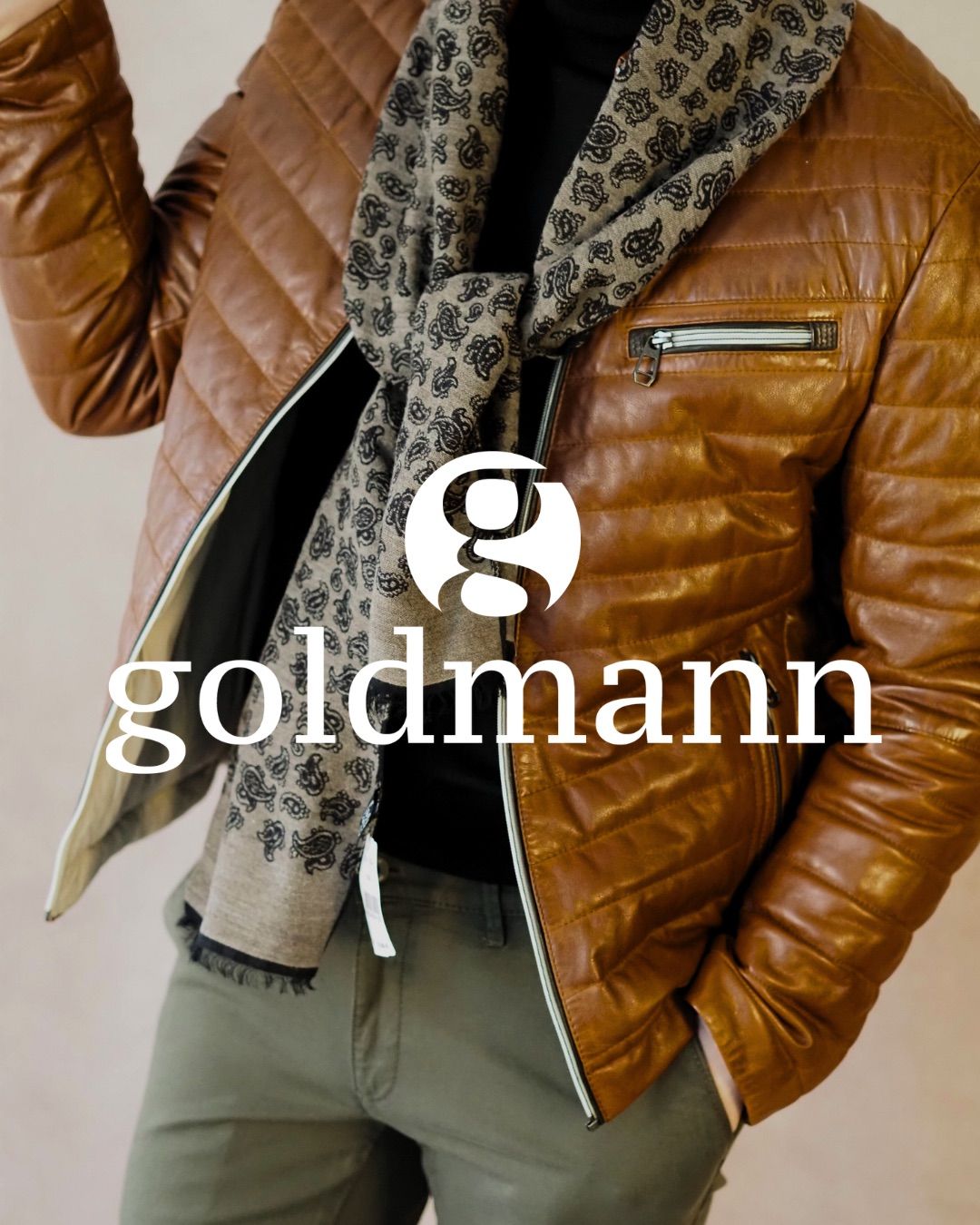 Ein Mann trägt eine braune Lederjacke und einen gemusterten Schal. Die Jacke hat Reißverschlüsse und Taschen. Unten steht das Wort 'Goldmann'.