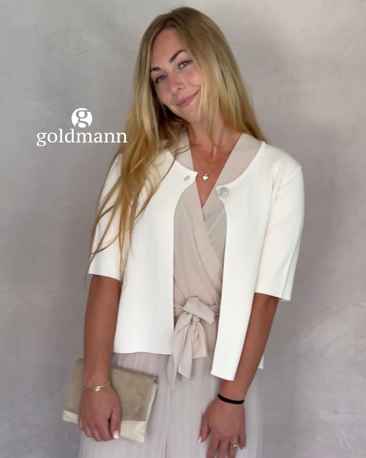 A woman with blonde hair is wearing a white blazer and a beige dress, smiling and holding a beige clutch. The image has a watermark of the brand 'goldmann'.