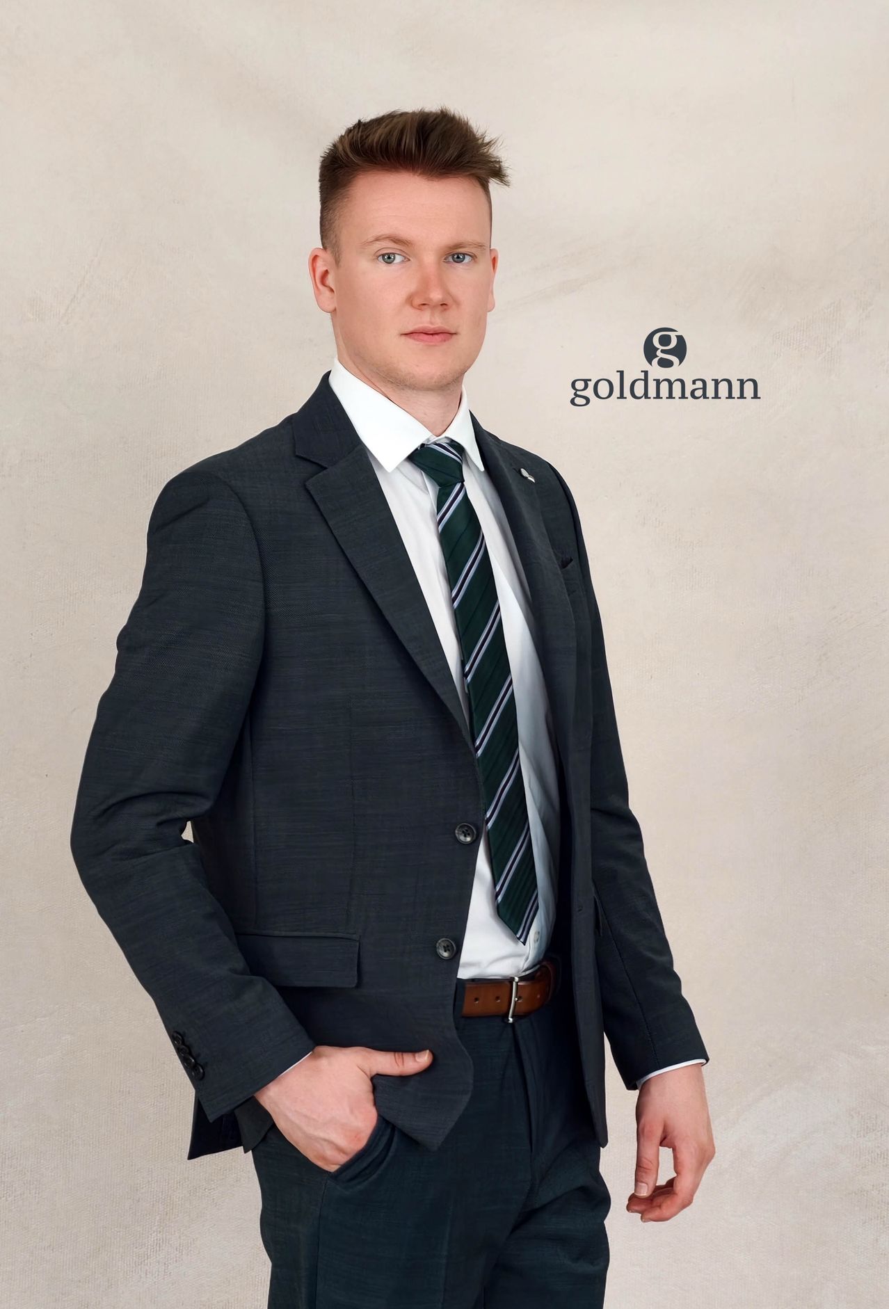 A man in a dark suit and tie, standing with a slight smile and one hand in his pocket. The background is a plain, light-colored wall.