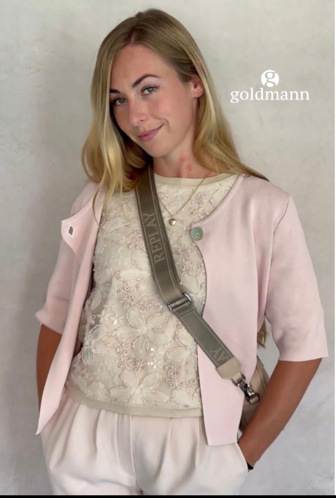 Eine Frau in einer rosa Jacke und einem weißen Kleid mit Blumenmustern, das eine Replay-Umhängetasche trägt. Sie hat blondes Haar und lächelt.