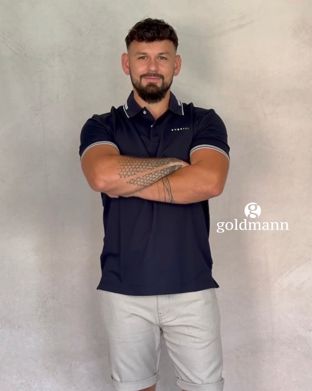 A man with a beard and tattoos on his arms is standing with his arms crossed wearing a navy blue polo shirt.