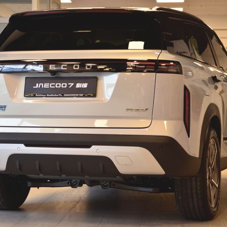 Ein weißer SUV mit dem Kennzeichen JNECOO7 SHS steht in einem Ausstellungsraum. Er hat ein schlankes Design mit LED-Rückleuchten und einem glänzenden schwarzen Stoßfänger.
