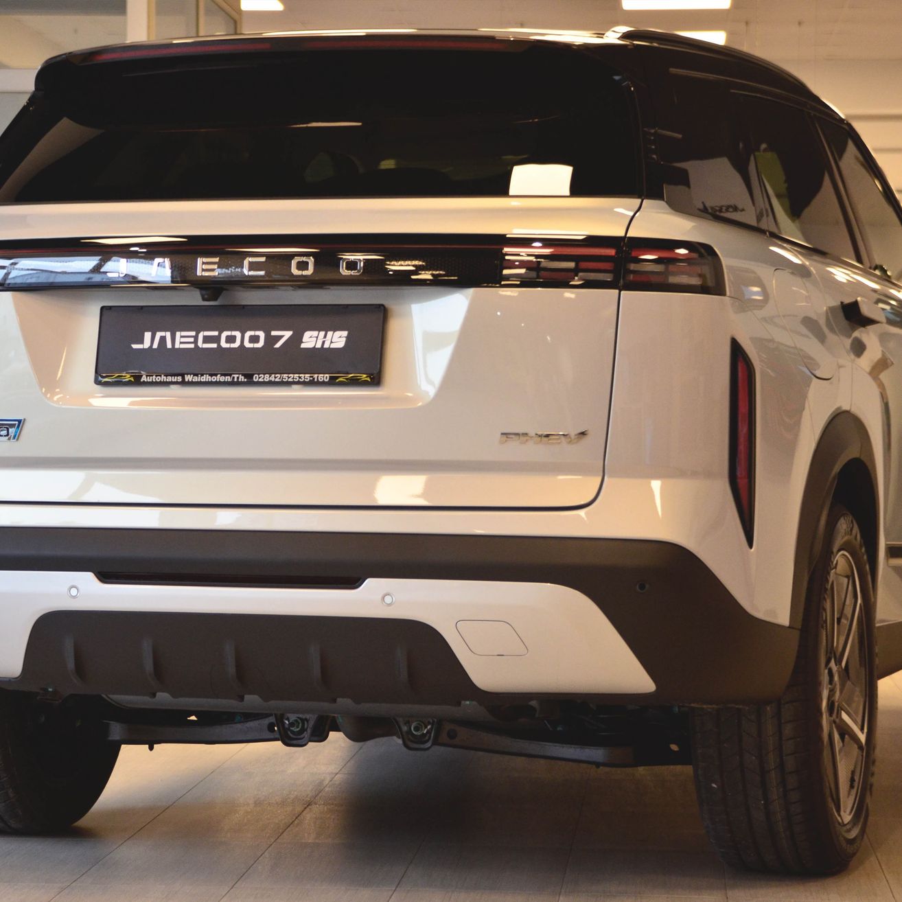Ein weißer SUV mit dem Kennzeichen JNECOO7 SHS steht in einem Ausstellungsraum. Er hat ein schlankes Design mit LED-Rückleuchten und einem glänzenden schwarzen Stoßfänger.