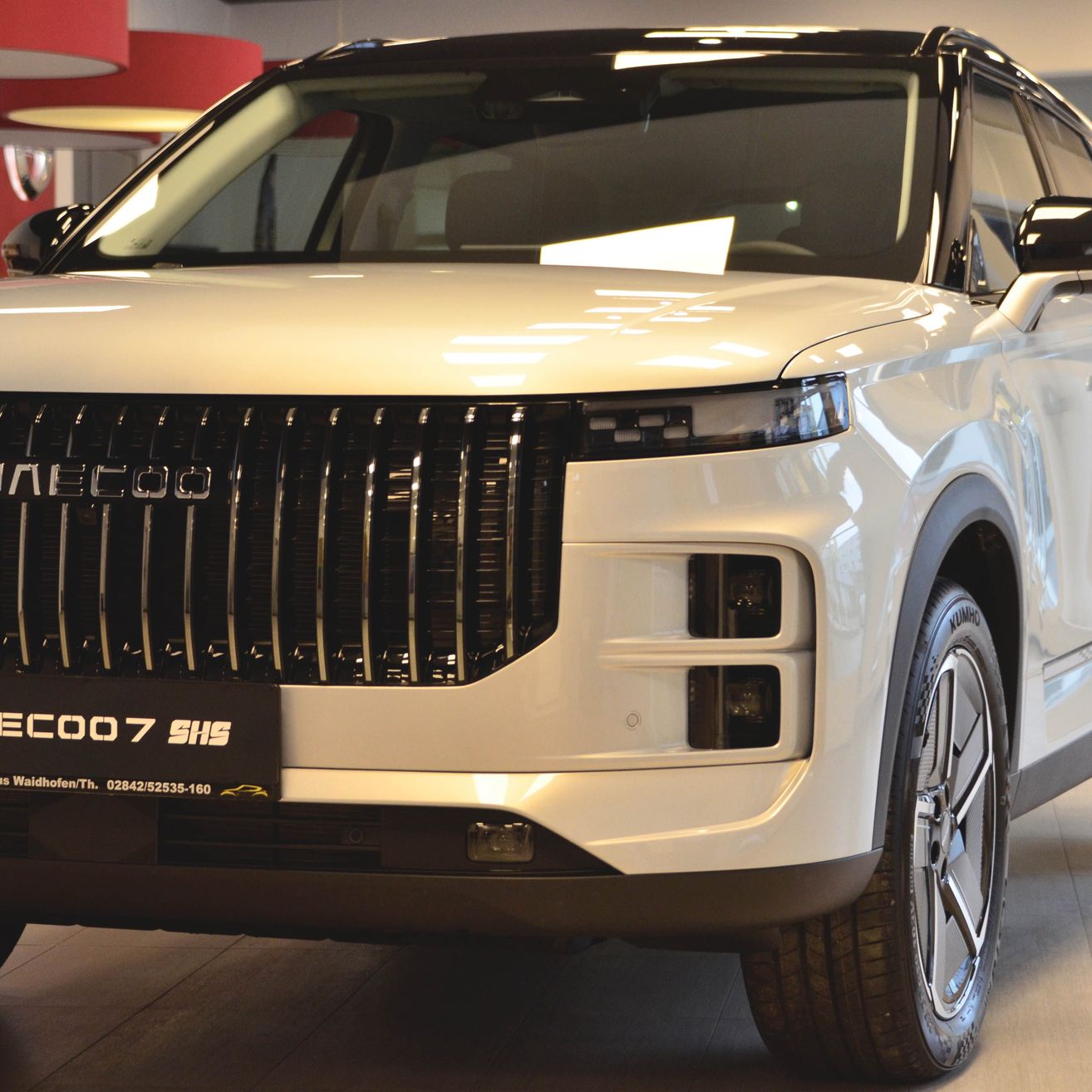 Ein weißes Fahrzeug mit dem Modellnamen MEECOO 7 SHS steht in einem Ausstellungsraum. Das Fahrzeug zeichnet sich durch ein schlankes Design mit schwarzem Frontgrill, LED-Scheinwerfern und einer glänzenden Außenseite aus.