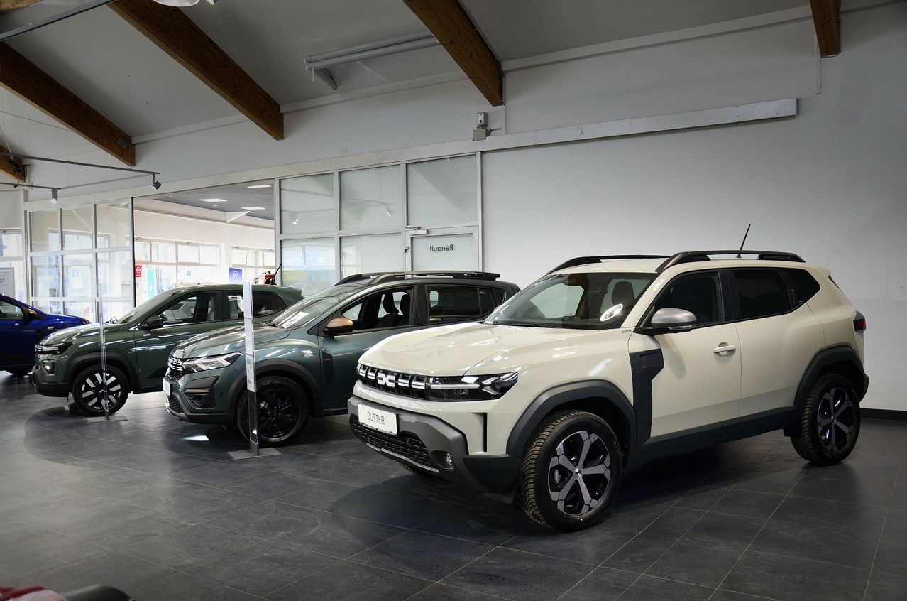 Ein Showroom mit mehreren Autos, darunter ein weißes Auto mit dem Wort 'Duster' auf dem Nummernschild, wird auf einem schwarzen Fliesenboden ausgestellt.