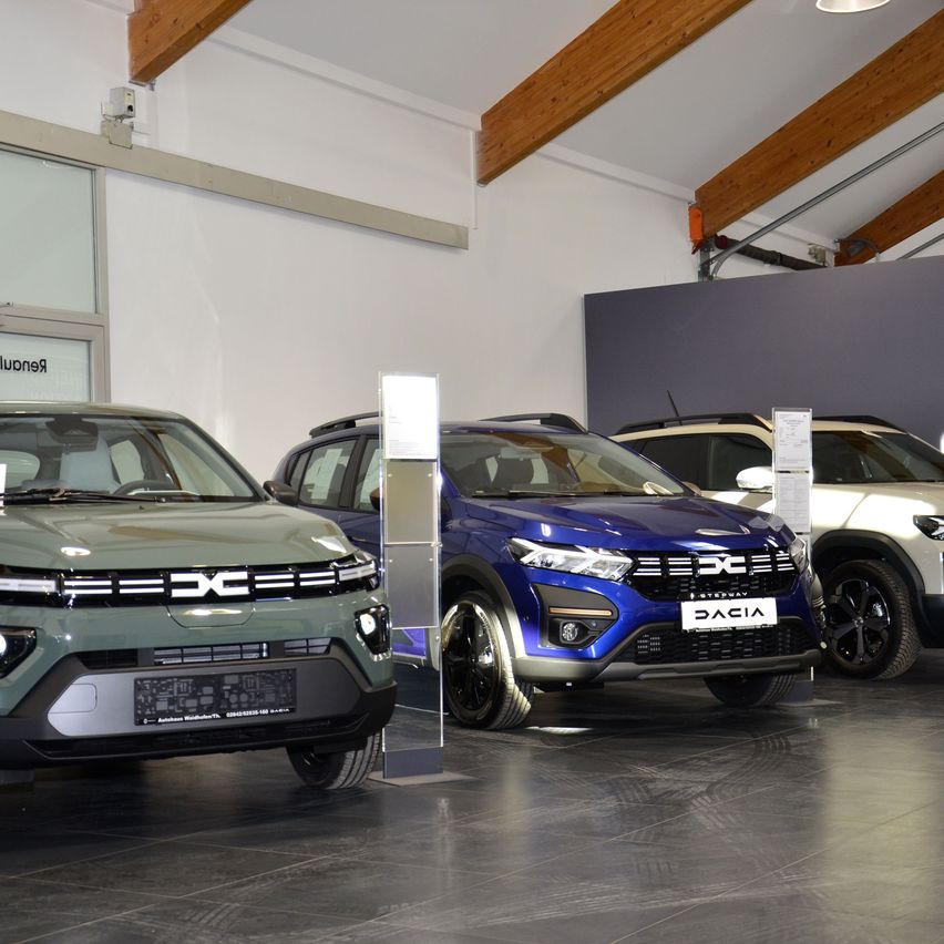 Drei Dacia-Fahrzeuge sind in einem Ausstellungsraum geparkt und zeigen ihre charakteristischen Kühlergrills und vorderen Stößstangen. Der Ausstellungsraum hat eine Holzdecke und weiße Wände.