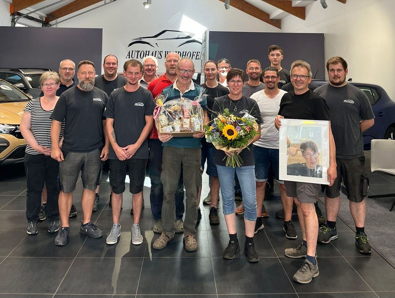 Eine Gruppe von Menschen steht in einem Raum mit gefliestem Boden und lächelt für ein Foto. Einige tragen Brillen, und eine Person hält einen Blumenstrauß und einen Geschenkkorb. Ein Schild steht Autohaus Undorfhof.
