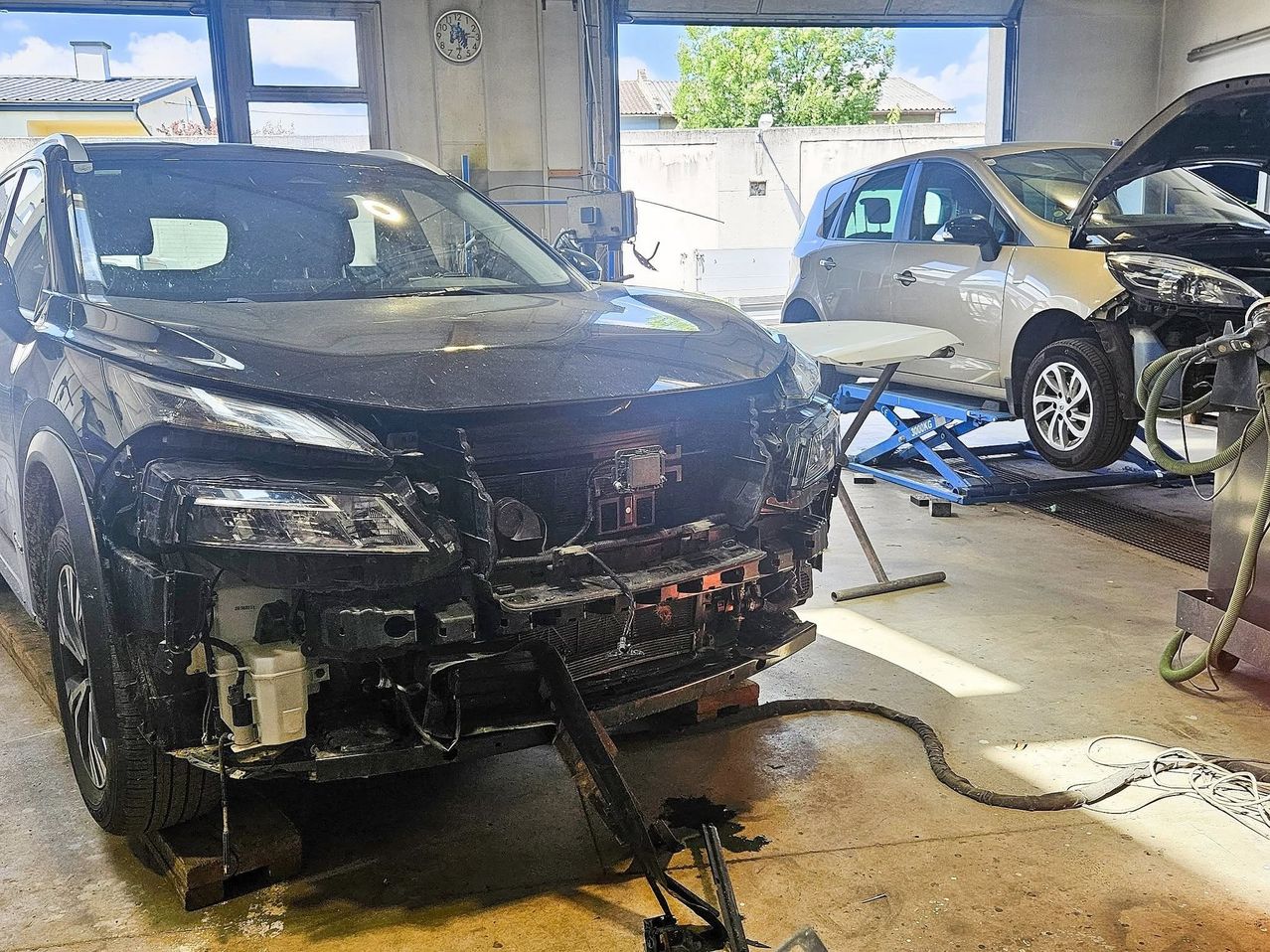 Ein schwarzer Wagen wird in einer Werkstatt bearbeitet. Der vordere Stoßfänger ist abmontiert. Im Hintergrund steht ein weißer Wagen auf einem Wagenheber.