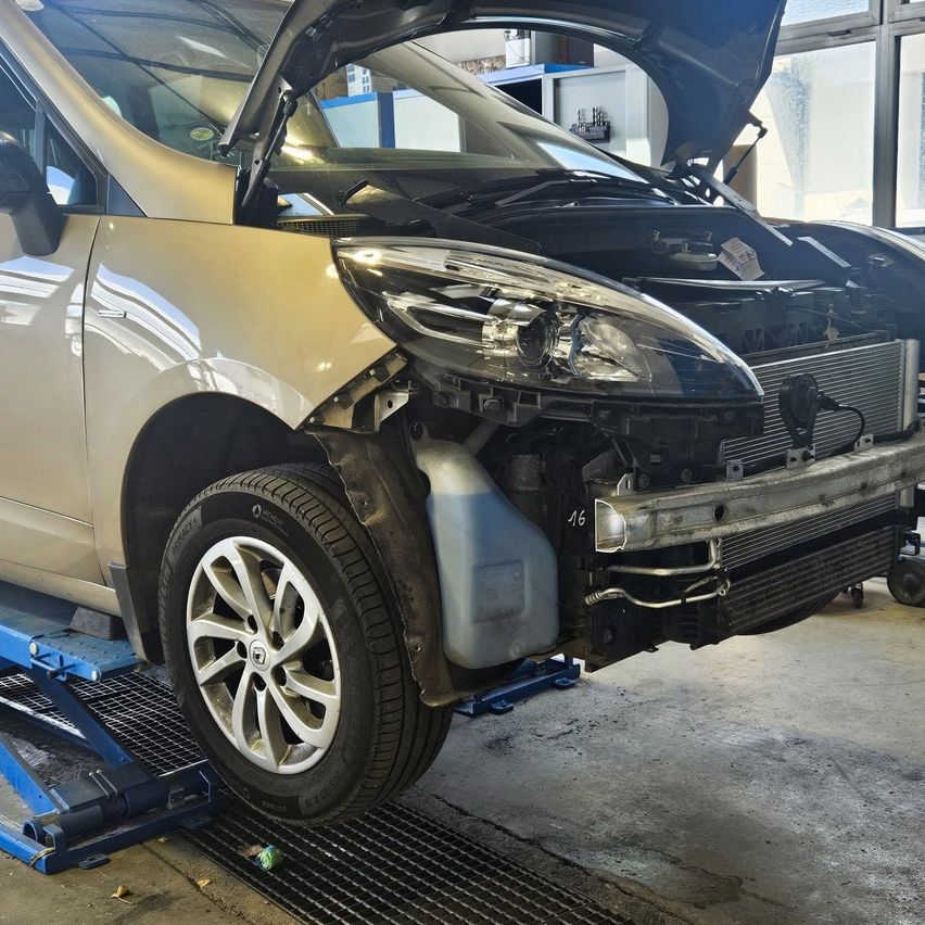 Ein Fahrzeug ist auf einem Wagenheber angehoben und zeigt die Vorderseite mit geöffneter Motorhaube, die den Motor enthüllt. Der Stoßfänger, der Kühler und ein Teil des Motorraums sind sichtbar.