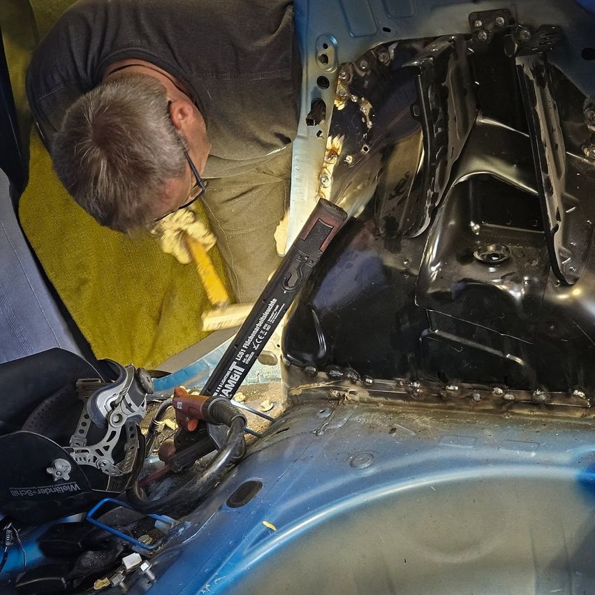 Ein älterer Mann mit Brille arbeitet am Motorraum eines Fahrzeugs und verwendet ein Werkzeug, um Komponenten zu justieren oder zu überprüfen. Das Auto ist zerlegt und zeigt Metallteile und Verkabelung.