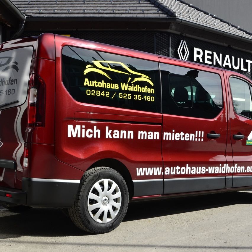 Ein roter Renault-Transporter ist vor einem Gebäude mit dem Renault-Logo geparkt. Er trägt die Aufschrift 'Autohaus Waidhofen' und eine Telefonnummer an der Seite. Der Transporter hat auch eine Website an der Seite geschrieben.