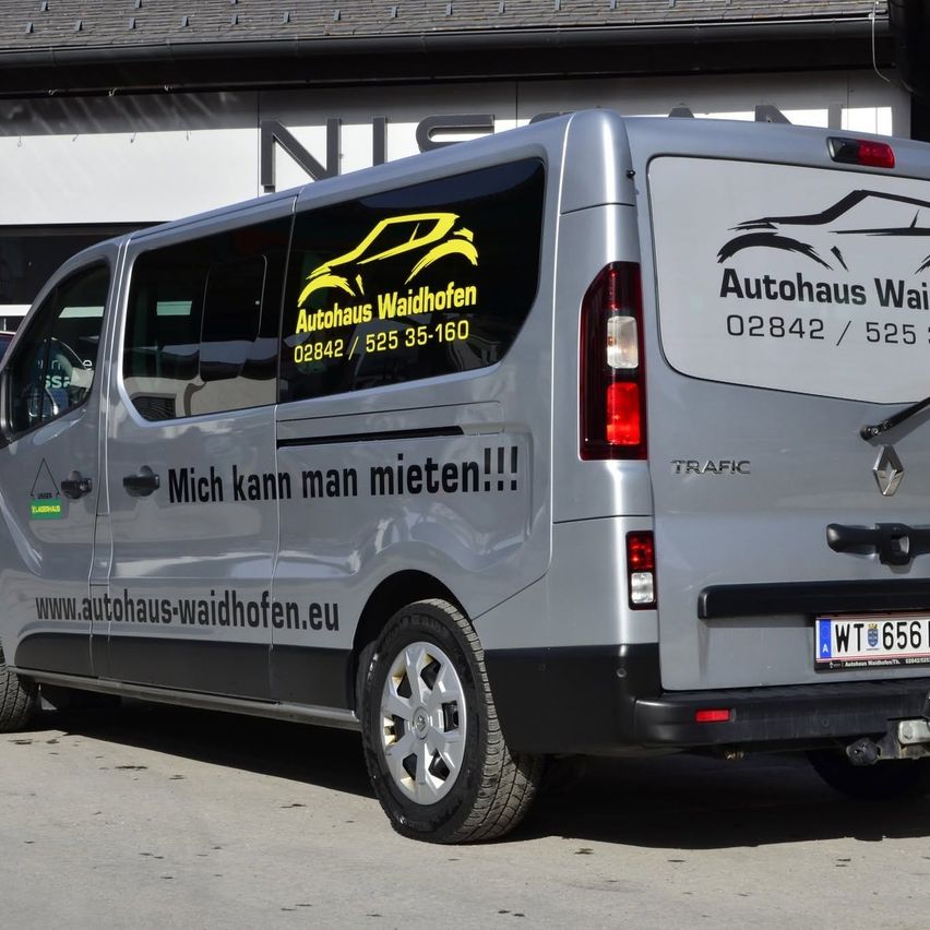 Ein silberner Van mit 'Autohaus Waidhofen' auf der Seite, mit einem gelben Auto-Logo und dem Text 'Mich kann man mieten!!!!' in schwarz. Die Rückseite des Vans hat eine Telefonnummer und eine Website-Adresse.