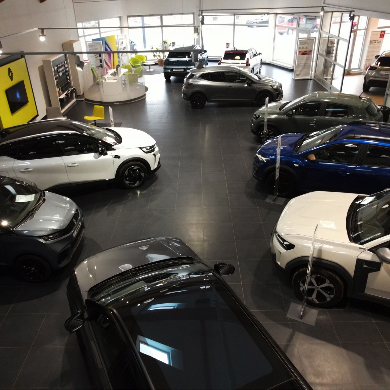 Ein Autohaus zeigt verschiedene Autos in verschiedenen Farben und Modellen. Die Autos sind in einer Reihe geparkt mit ausreichend Platz zwischen ihnen. Das Showraum hat ein modernes Design mit gefliestem Boden und Glaswänden.