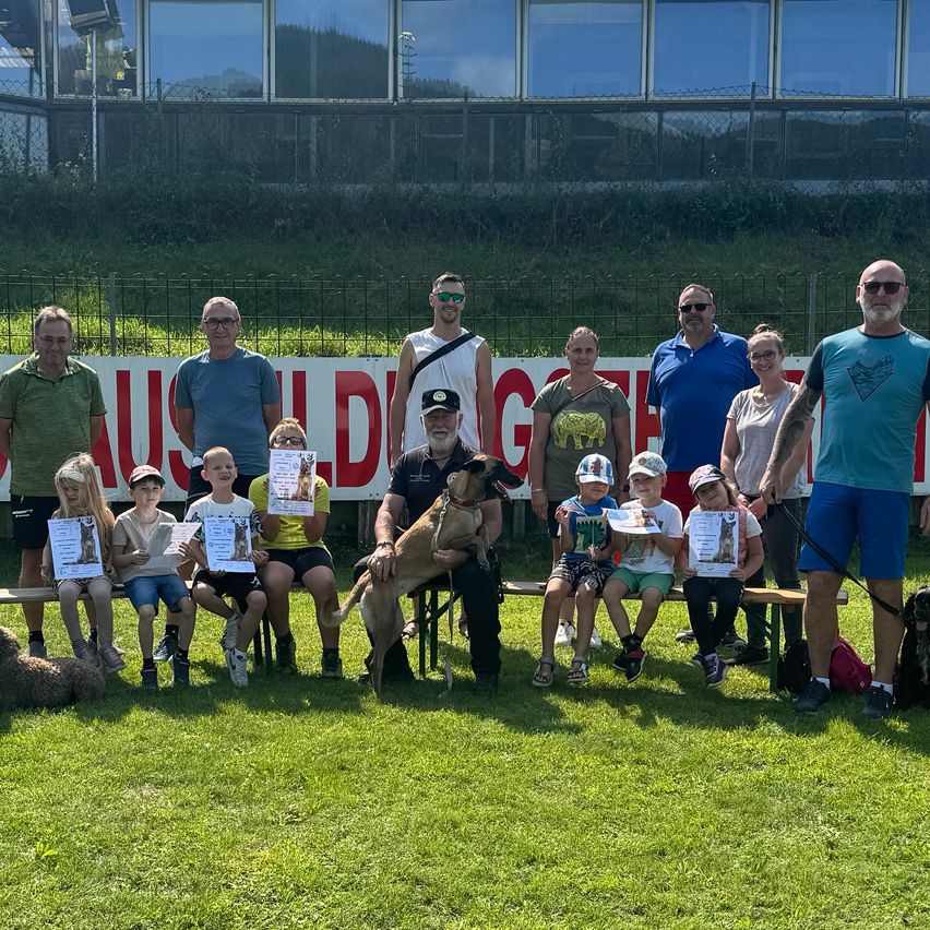 Eine Gruppe von Erwachsenen und Kindern posiert mit Hunden auf einem Rasenfeld. Sie halten Urkunden in der Hand und sitzen auf einer Bank mit Hunden um sie herum.