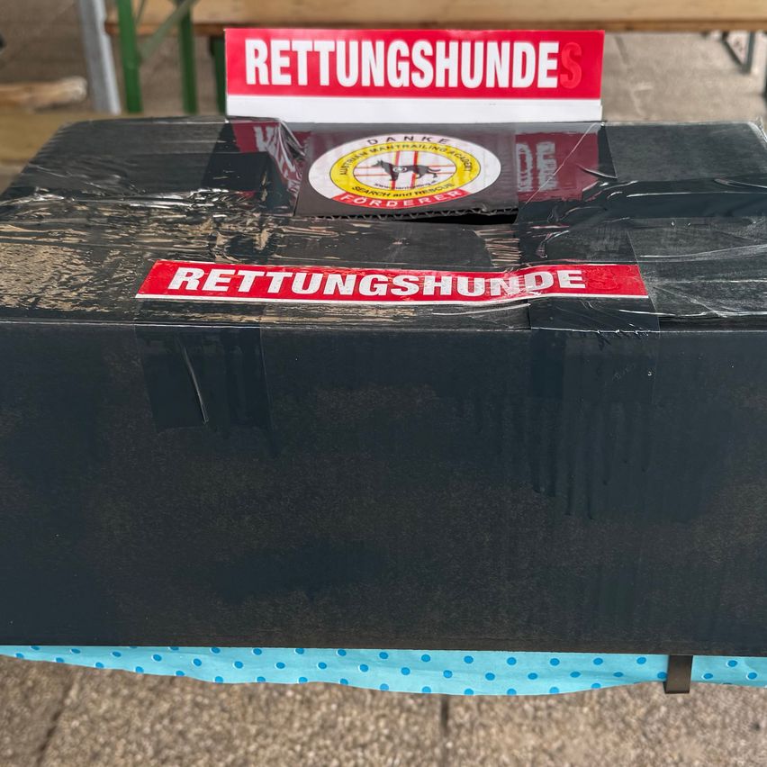 Eine schwarze Box mit Plastik- und blau gepunktetem Stoff, mit rotem Text 'Rettungshunde'. Oben befindet sich ein rotes und weißes Schild mit demselben Text.