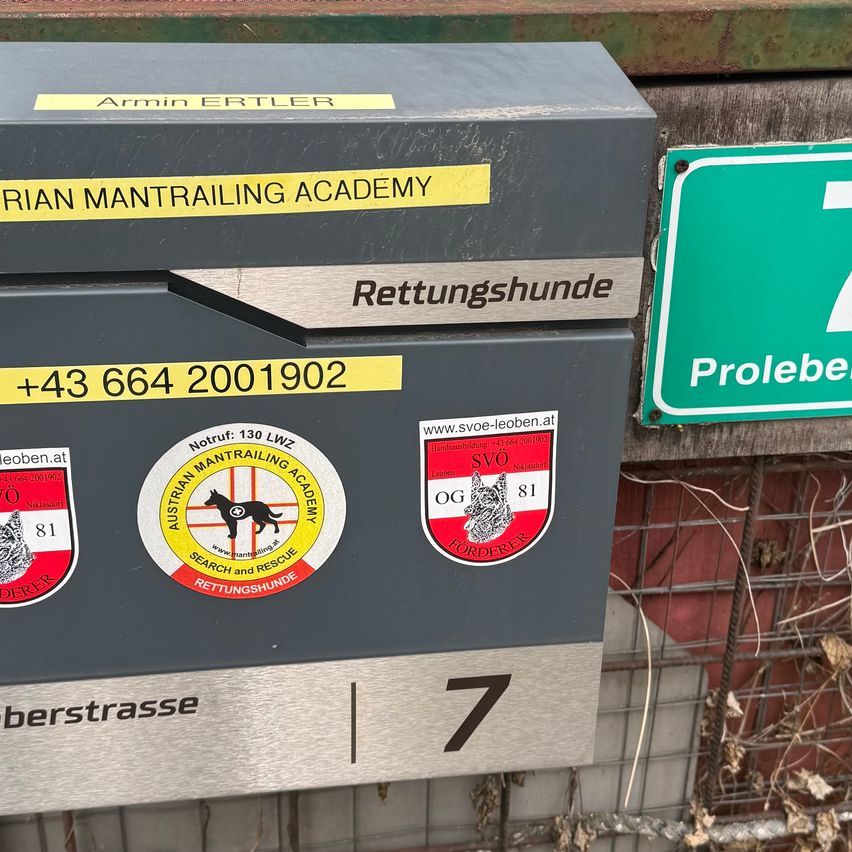 Eine graue Aufbewahrungsbox mit gelben Aufklebern trägt die Aufschrift 'Arian Mantrailing Academy' und 'Rettungshunde'. Sie enthält Logos und Telefonnummern. Ein grünes Schild mit weißen Buchstaben steht 'Prolebe'. Dahinter befindet sich ein Metallzaun.