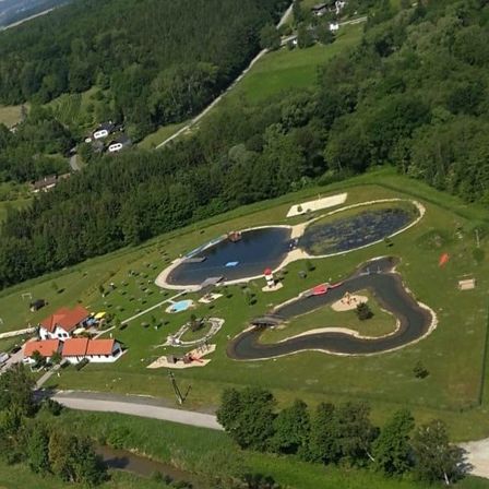 Ein Luftbild einer großen Außenrecreationsfläche mit einem See, einer roten Rutsche, einer Rennstrecke und Häusern. Bäume und Pflanzen umgeben das Gebiet.