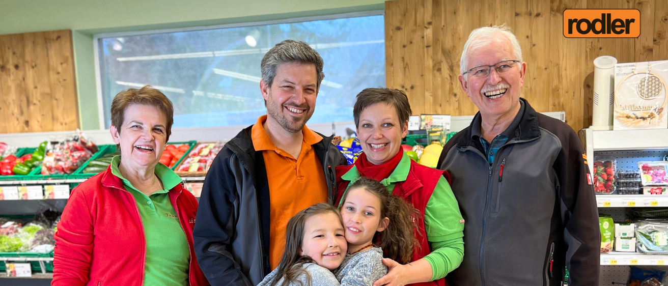 Eine Familie mit vier Erwachsenen und zwei Kindern steht zusammen in einem Geschäft. Sie alle lächeln und posieren für ein Foto. Sie stehen vor einem Regal mit verschiedenen Produkten.