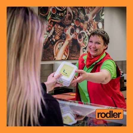 Eine lächelnde Mitarbeiterin übergibt einem Kunden einen Abholbehälter in einem Restaurant mit einem farbenfrohen Wandbild im Hintergrund.