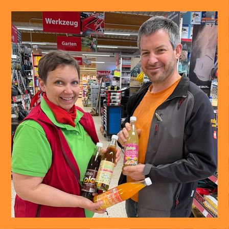Zwei Menschen lächeln in einem Geschäft. Die Frau hält zwei Flaschen Saft, der Mann eine. Beide tragen Freizeitkleidung.