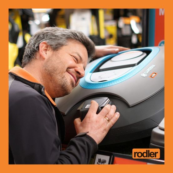 Ein Mann lächelt, während er ein graues und blaues Gerät mit der Aufschrift 'Garmin' in einem Geschäft hält. Er trägt einen schwarzen Mantel und einen Ring.