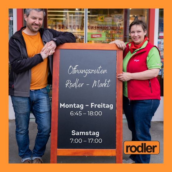 Zwei Personen stehen neben einem schwarzen Brett, das die Öffnungszeiten des Geschäfts anzeigt. Ein Mann und eine Frau lächeln und stehen vor einem Geschäft.