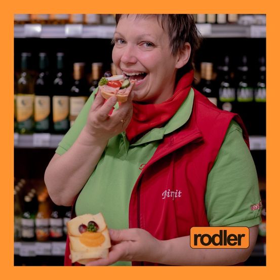 Eine Frau in einem grünen Hemd und einer roten Weste isst in einem Supermarkt ein Sandwich und lächelt. Sie hält ein weiteres Sandwich in der Hand. Hinter ihr sind Regale mit Weinflaschen zu sehen.