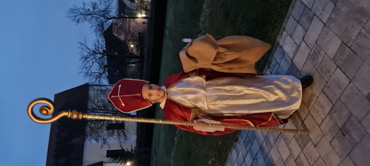 Ein junges Kind, verkleidet als Bischof, steht nachts in einem Grasbereich und hält einen Stab in der Hand. Gebäude sind im Hintergrund zu sehen.