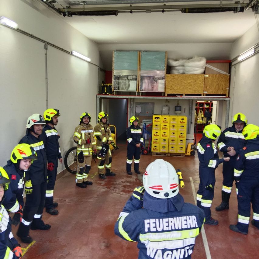 Eine Gruppe von Feuerwehrleuten in voller Ausrüstung, Helmen und Schutzanzügen versammelt sich in einer Feuerwache. Im Hintergrund sind Ausrüstungsgegenstände und Fahrräder zu sehen.