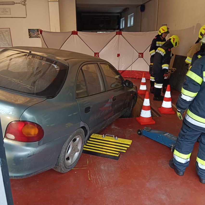 Ein graues Auto ist in einer Garage auf einem Wagenheber gehoben und von Verkehrskegeln umgeben. Feuerwehrleute in gelben und schwarzen Uniformen sind in der Nähe, einer schaut in das Auto.