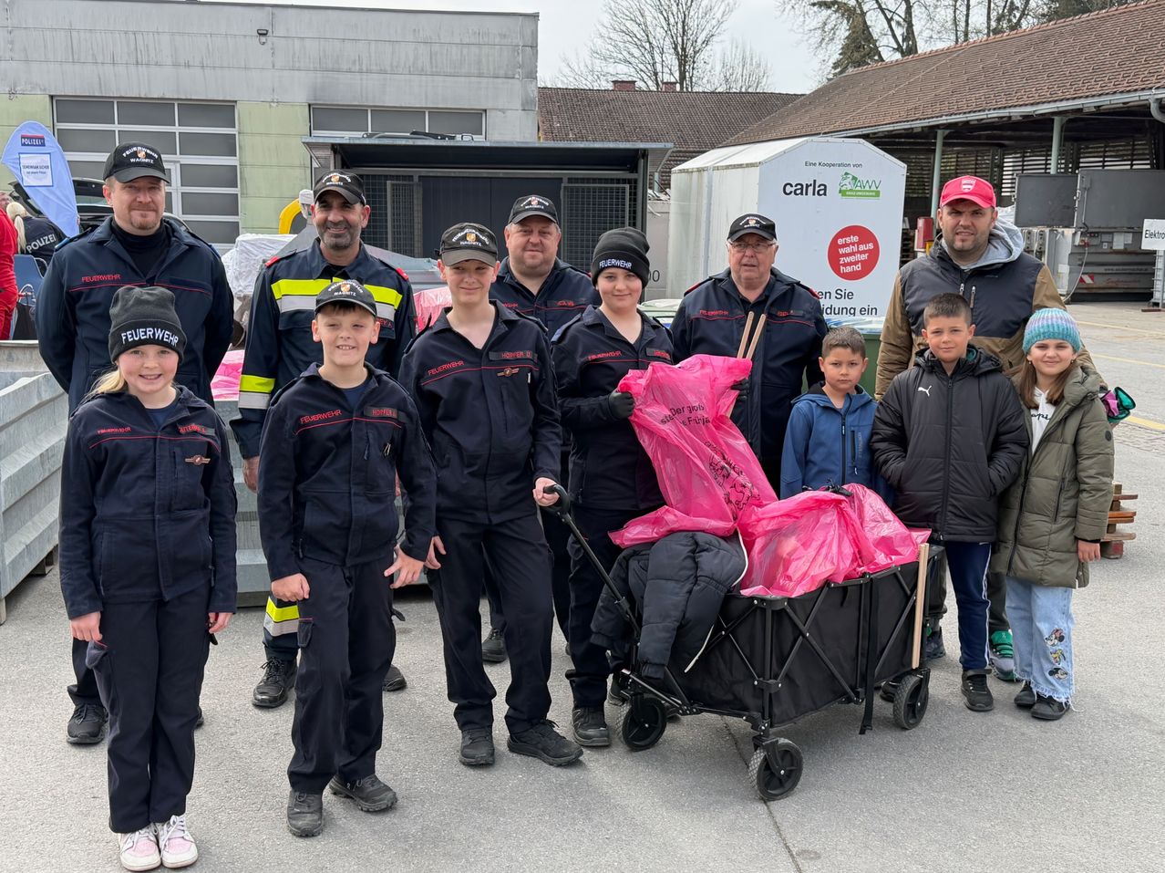 A group of firefighters and children stand outside, some holding pink bags. A stroller with pink bags is pushed by a person. Buildings and trees are in the background.